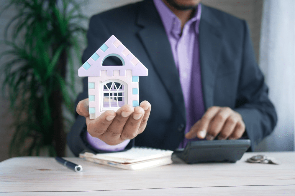 A Faceless Male in a Suit Holding out a Miniature Purple Home While Using a Calculator with the Other 