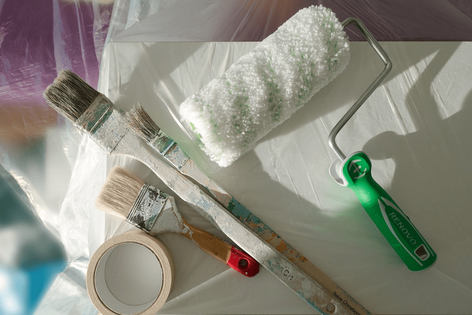 A Slightly Tinged Paint Roller, Three Brushes, and A Tape Resting on a Plastic-Covered Surface