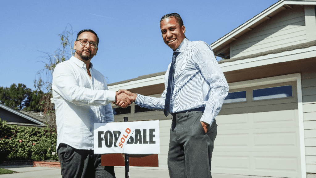 Two men shaking hands in front of a house with a for sale sign.