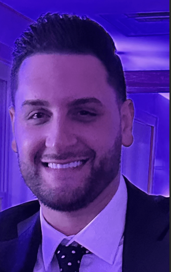 A man in a suit and tie smiling in front of a purple light.