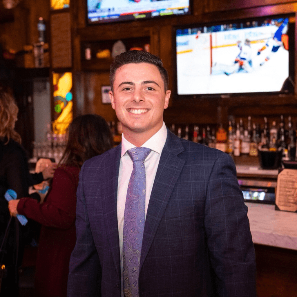 A smiling man in a suit and tie at a social event with a bar and a television in the background.