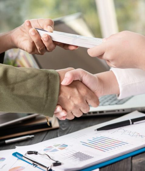 Two individuals shaking hands over a desk while exchanging business cards.