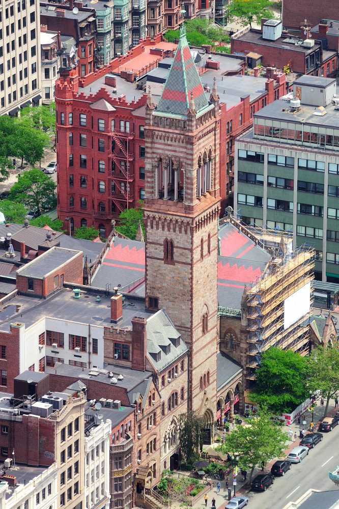 Aerial view of a historic red brick church with a green spire among modern urban buildings.