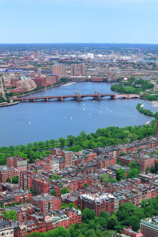 Aerial view of a city with dense brick buildings, a bridge crossing over a river with boats, and greenery in the foreground.