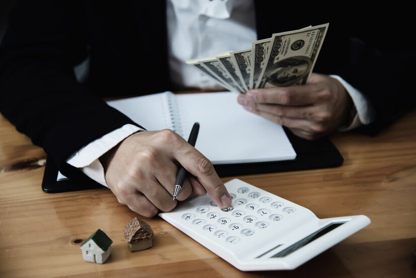 A person calculating finances with cash in hand, a calculator, and miniature houses on the table.