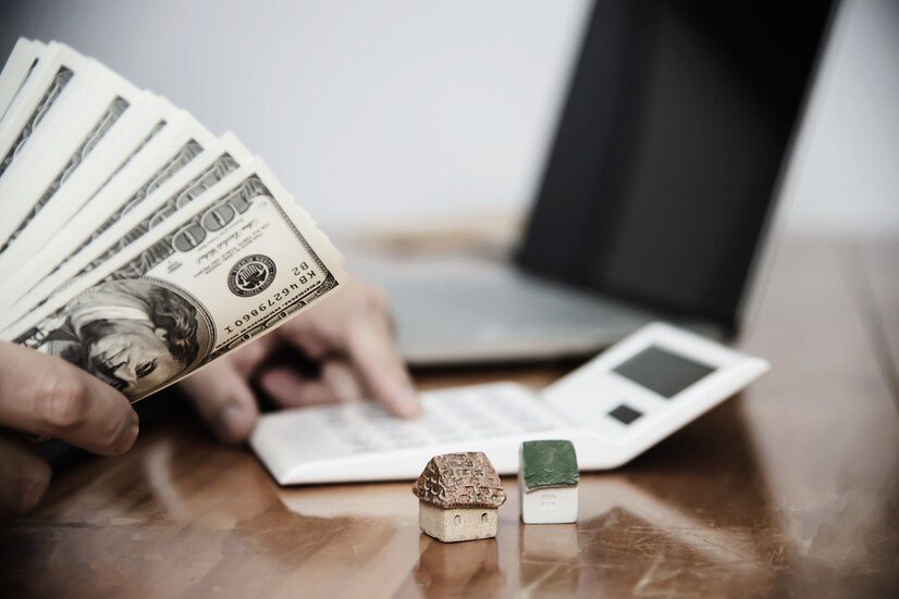 Person calculating finances or budget with a stack of cash and miniature houses on the desk, suggesting real estate or mortgage planning.