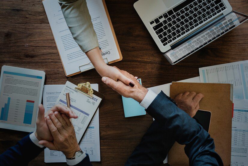 Two professionals shaking hands across a table with business documents and a laptop.