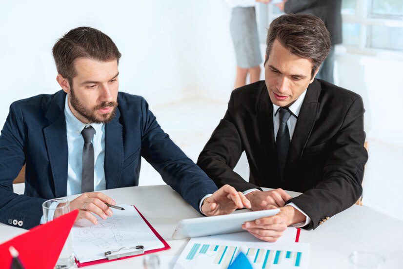 Two businessmen discussing over a digital tablet with reports on the table.