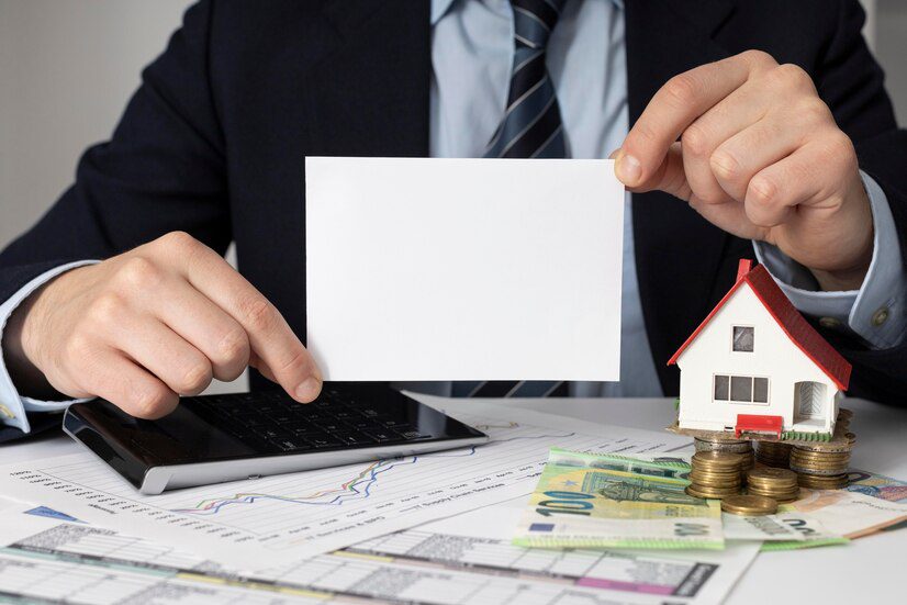 Professional presenting a blank card with a model house and coins on financial documents, symbolizing real estate investment or mortgage concept.