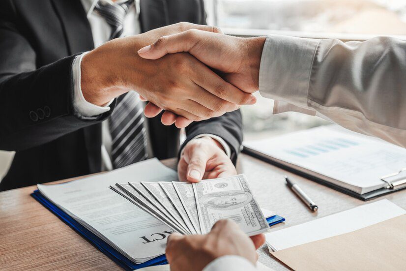 Two individuals shaking hands over a desk with money and documents, suggesting a financial agreement or deal.