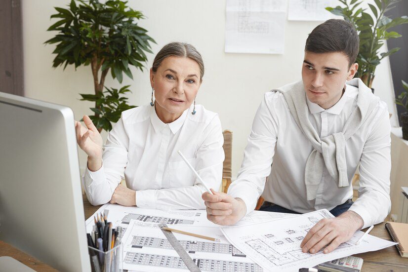 Two professionals reviewing documents at a desk with a computer and architectural plans.