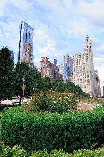 Urban park with greenery in the foreground and skyscrapers rising in the background against a cloudy sky.