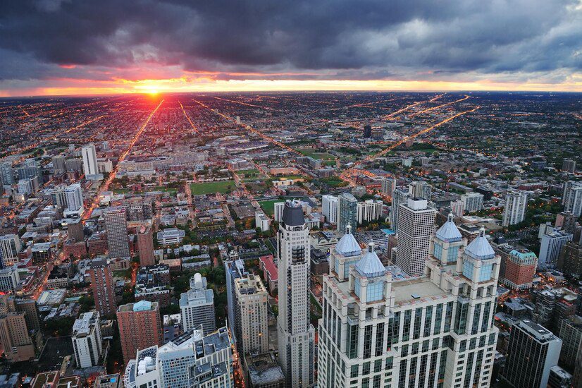 Aerial view of a cityscape at sunset with the sun casting a warm glow on the horizon and long shadows over the buildings and streets.