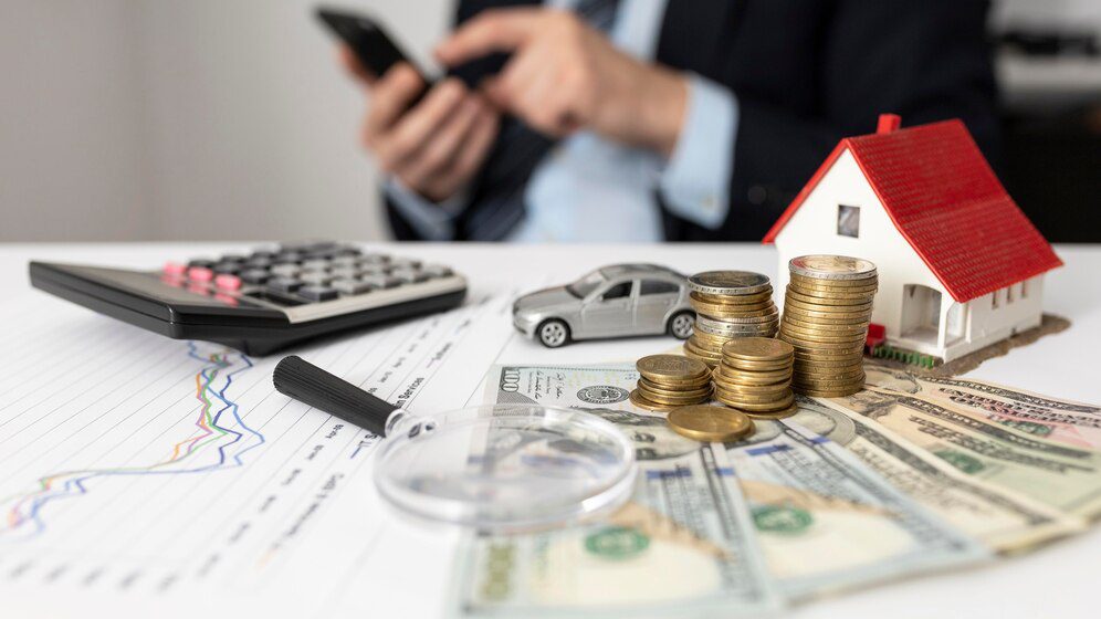 A person in a business suit managing finances with a calculator and smartphone, surrounded by a model house, a toy car, coins stacked, currency notes, and a graph analysis on a desk.