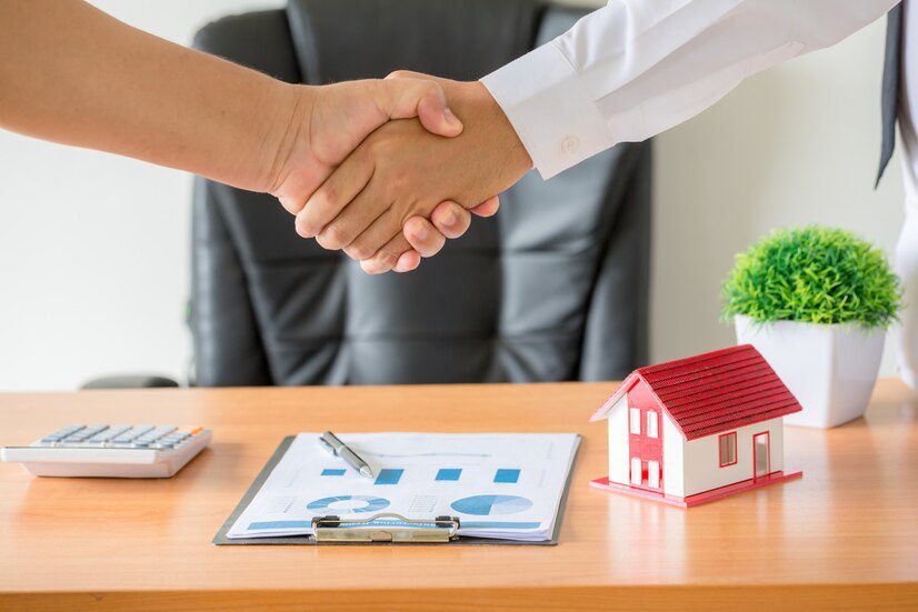 Two individuals shaking hands over a desk with real estate documents, a calculator, and a small house model, suggesting a completed property transaction.