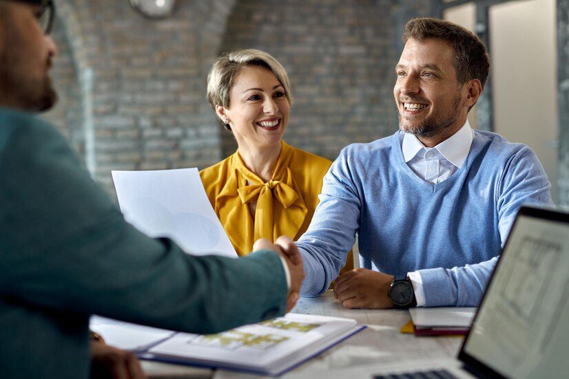 Two professionals shaking hands during a meeting, with a female colleague smiling in the background.