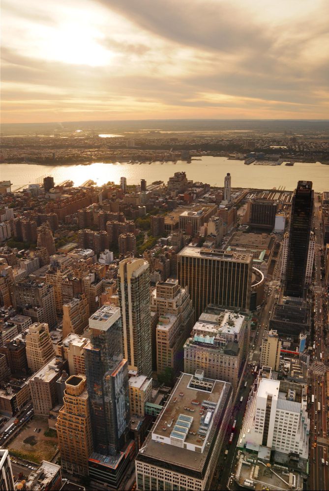 Aerial view of a densely built urban area with numerous skyscrapers at sunset, overlooking a wide river.