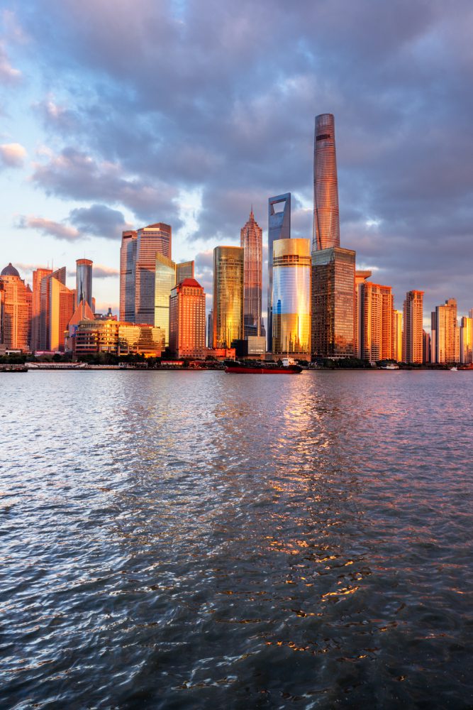 Sunset reflects on the skyline of a modern city with skyscrapers beside a calm body of water.