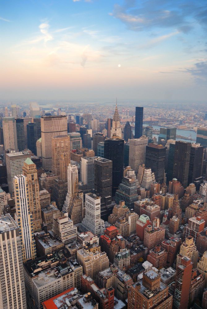 Aerial view of a densely built cityscape with skyscrapers at dusk, under a partially cloudy sky with a visible moon.