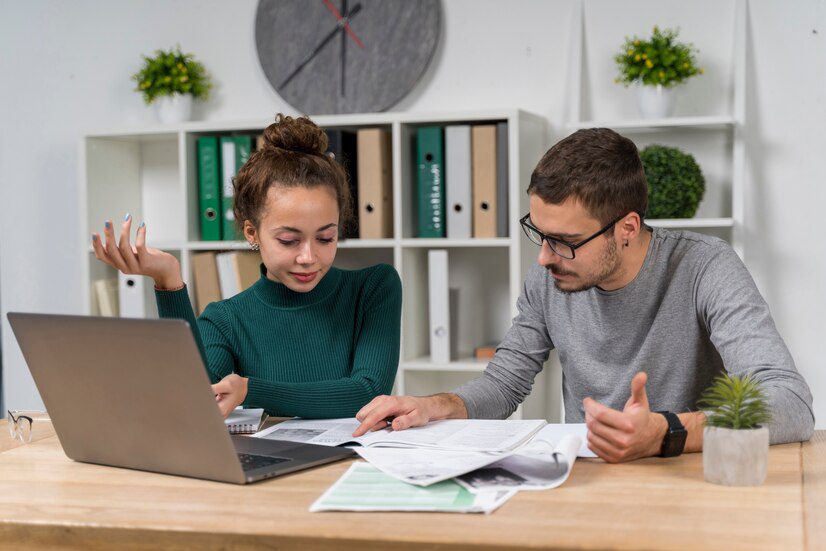 Two individuals reviewing documents and working on a laptop at an office desk.