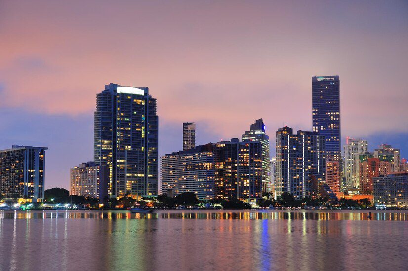 City skyline at dusk with illuminated buildings reflecting on the water.