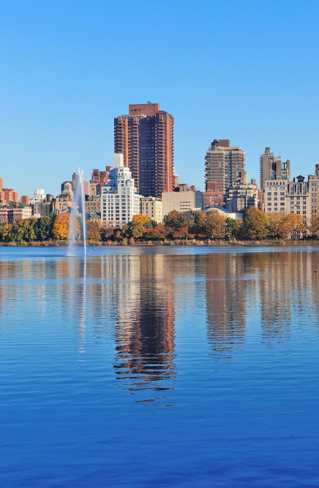 Skyline reflections on a calm water surface with a fountain in a city park.