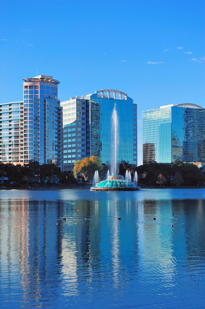 A cityscape featuring modern buildings reflected in a lake with a fountain.