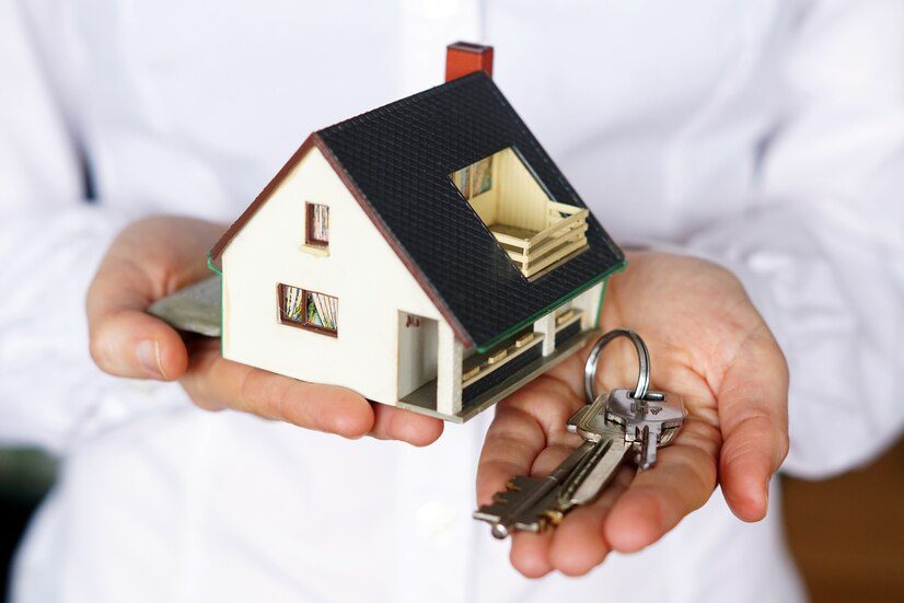 Person holding a small model house and a set of keys, symbolizing homeownership or real estate services.