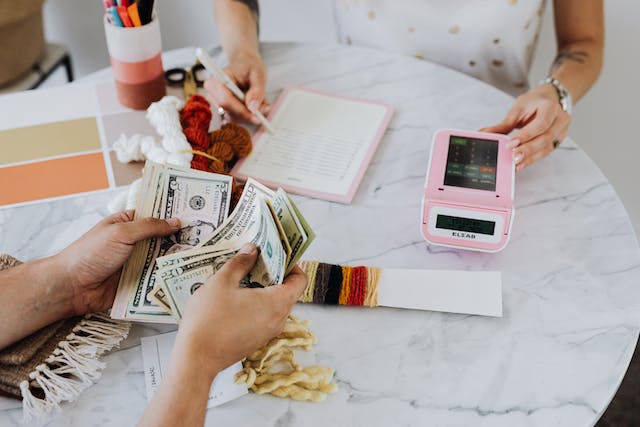 Two individuals at a table involved in a transaction with one person counting cash and the other recording details, alongside crafting materials and a pink calculator.