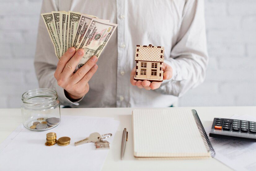 Person holding cash in one hand and a miniature house in the other over a desk with financial documents.