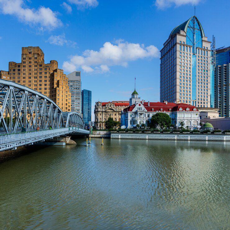 Steel truss bridge spanning a river with urban skyline and mix of modern and traditional buildings under a blue sky.