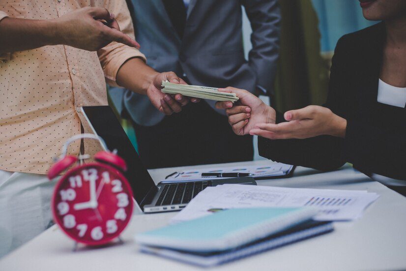 Persons exchanging money at a table with documents, a laptop, and a red clock present.