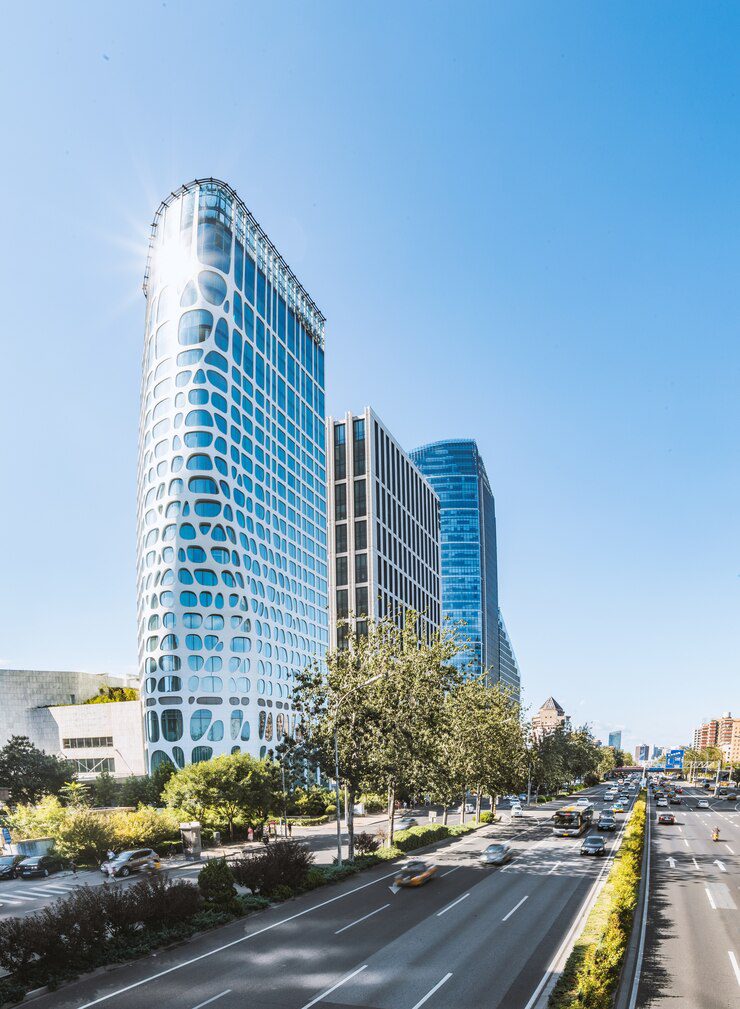 Modern skyscraper with a distinctive circular-patterned facade stands alongside a tree-lined urban road on a sunny day.