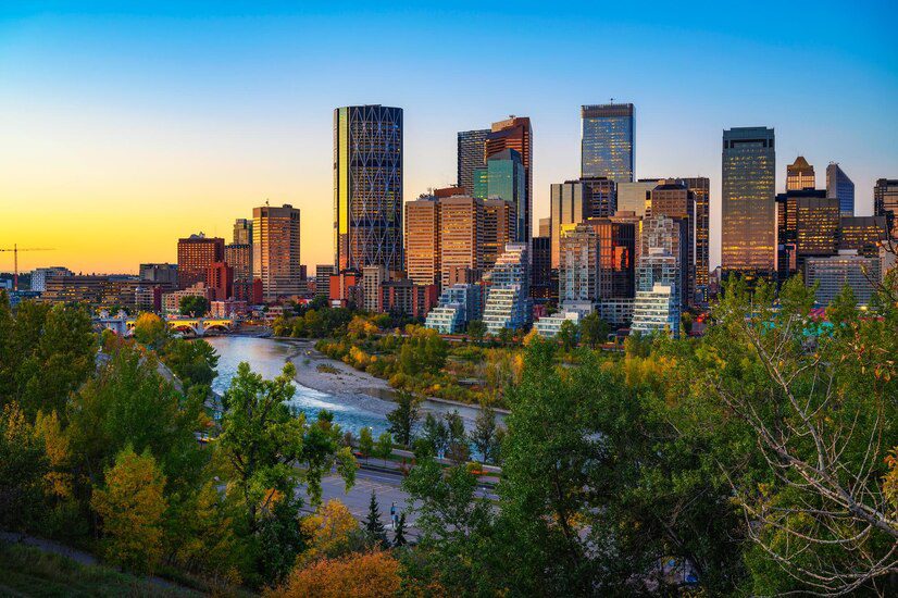 Skyline of a modern city at dusk with riverfront and autumnal trees.