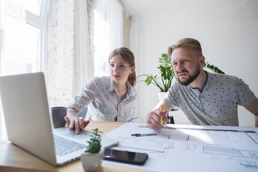 Two professionals working on a project with a laptop and architectural drawings.