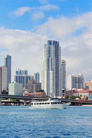 A yacht cruising along the waterfront with a backdrop of modern skyscrapers under a partly cloudy sky.