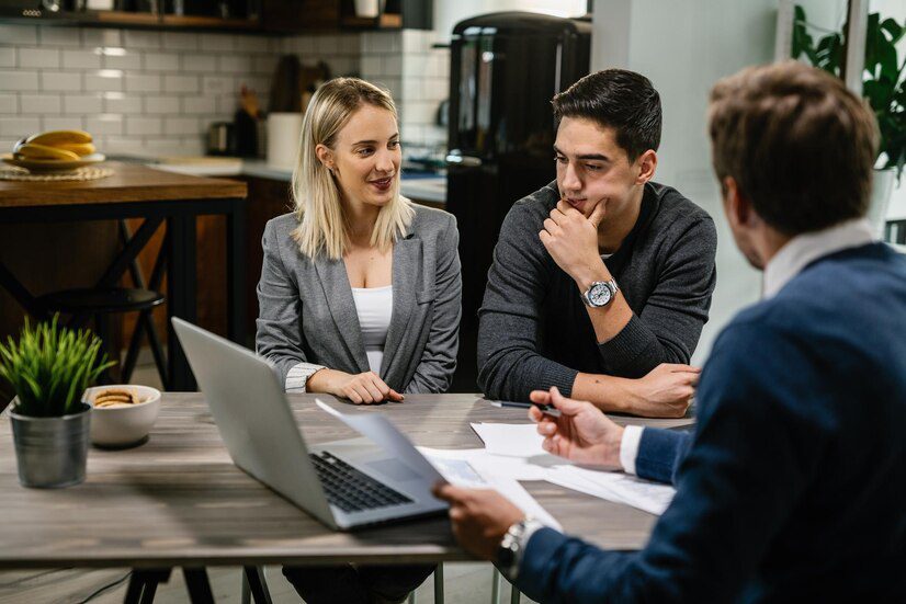 Three professionals engaged in a discussion around a meeting table with documents and a laptop.