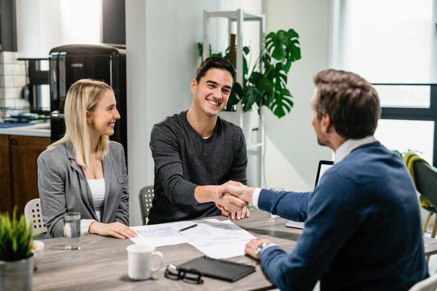 Two individuals smiling while one of them shakes hands with another person across the table during a meeting.