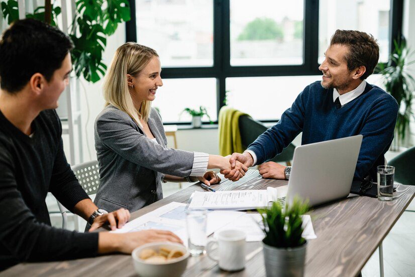 Two professionals shaking hands at a meeting table while another colleague observes.