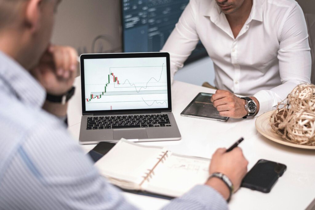 A man sits across from another man while reviewing a statistics chart on a laptop.