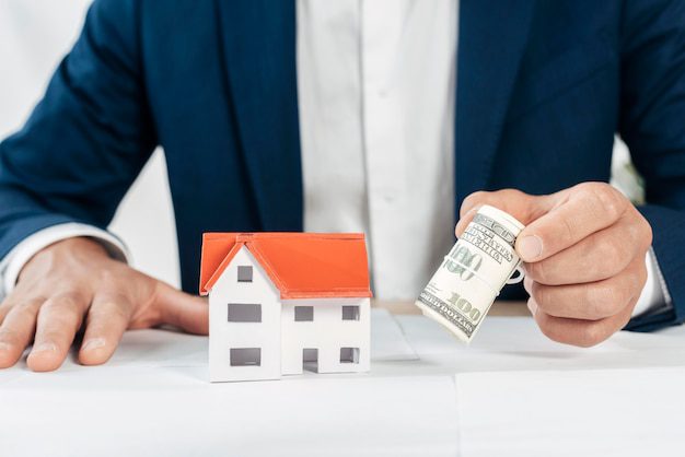 A person holding a roll of US dollar banknotes with a miniature house model on the table