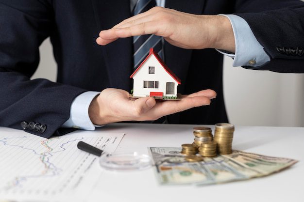 A person holding a miniature house model with coins, banknotes, and a magnifying glass on a table