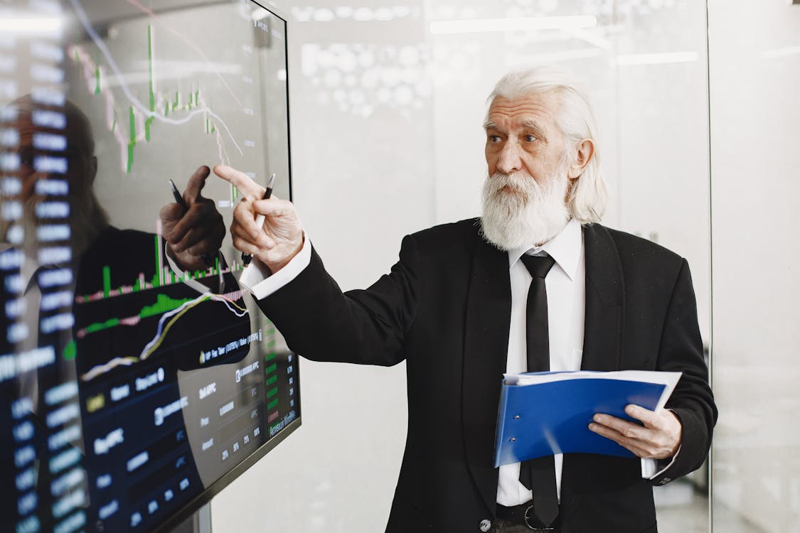 An elderly man points at charts on a screen, representing analysis for investment property mortgages.