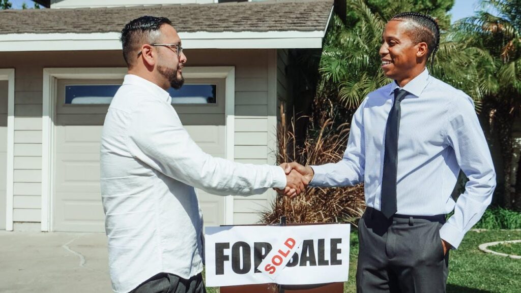 Two men shaking hands above a Sold sign in front of a house