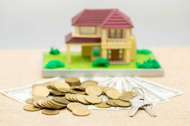 Dollars, coins, and a key on a surface in front of a miniature house model
