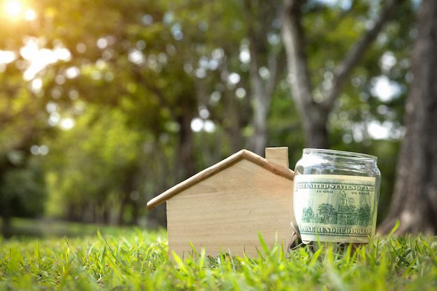 A jar with a dollar note beside a miniature wooden house on the grass