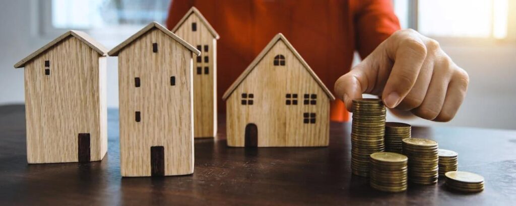 An image of four wooden houses on a table in front of a person with stacks of coins besides them.