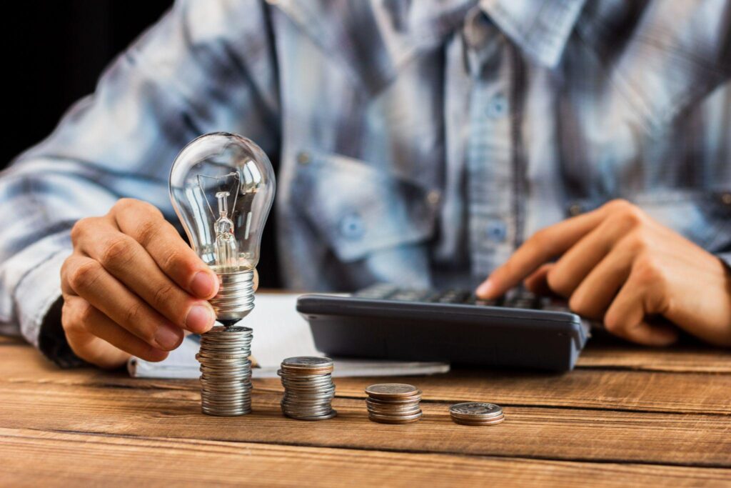 A light bulb on stacked coins with a man using a calculator in the background
