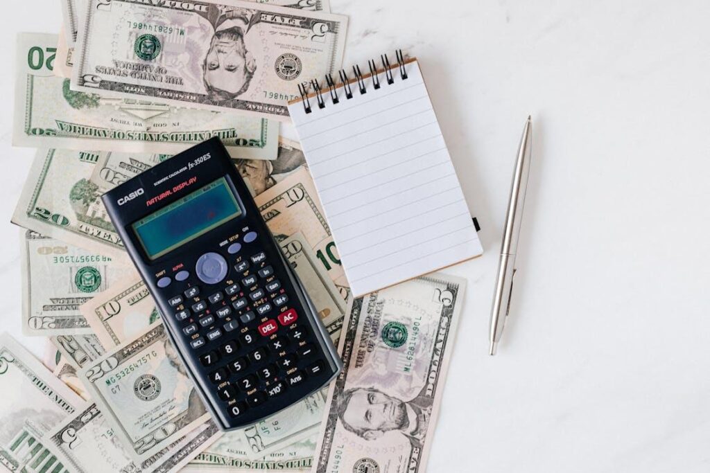 A flat lay image of scattered dollar bills with a calculator, note pad, and a pen lying on top.