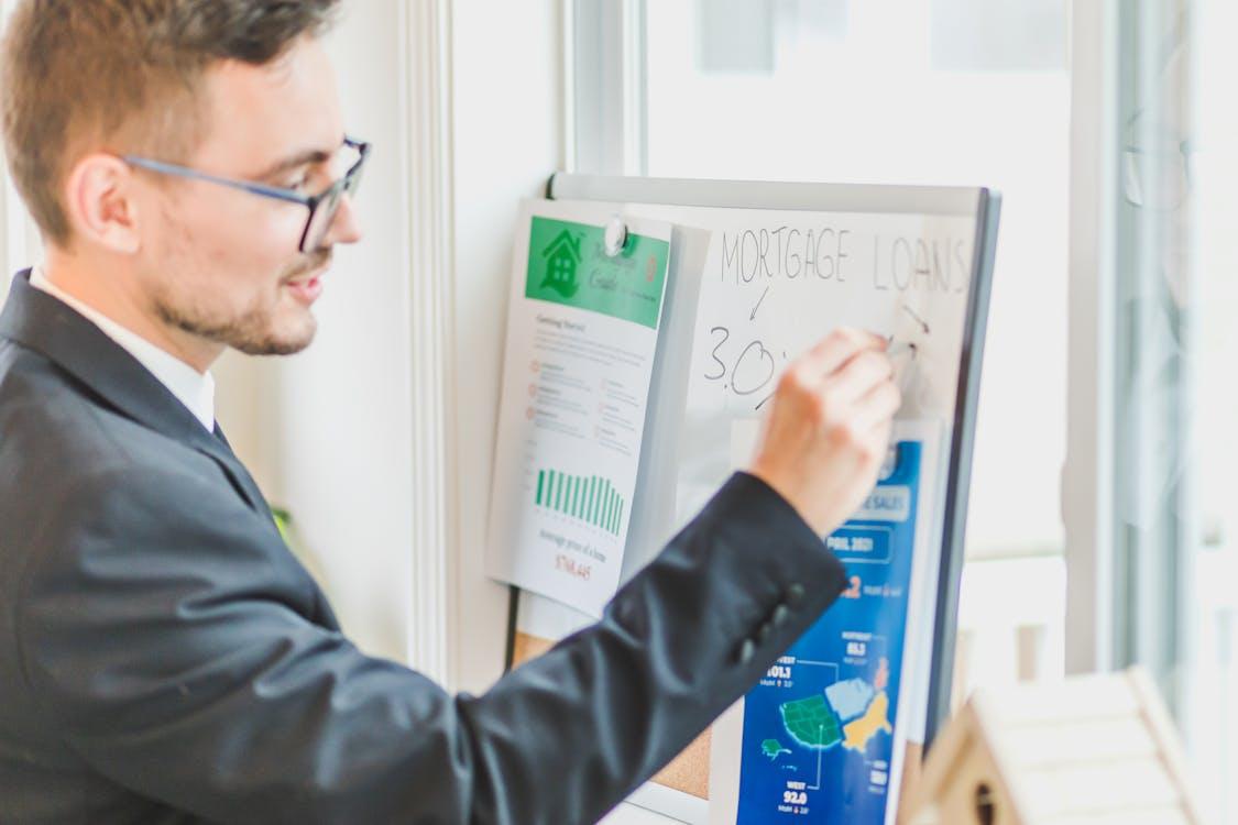 A man in a suit and glasses writes "Mortgage Loans" on a whiteboard